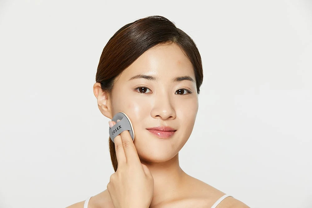 A woman with her hair pulled back uses a gray GIVEX facial cleansing device on her cheek against a clean white background.
